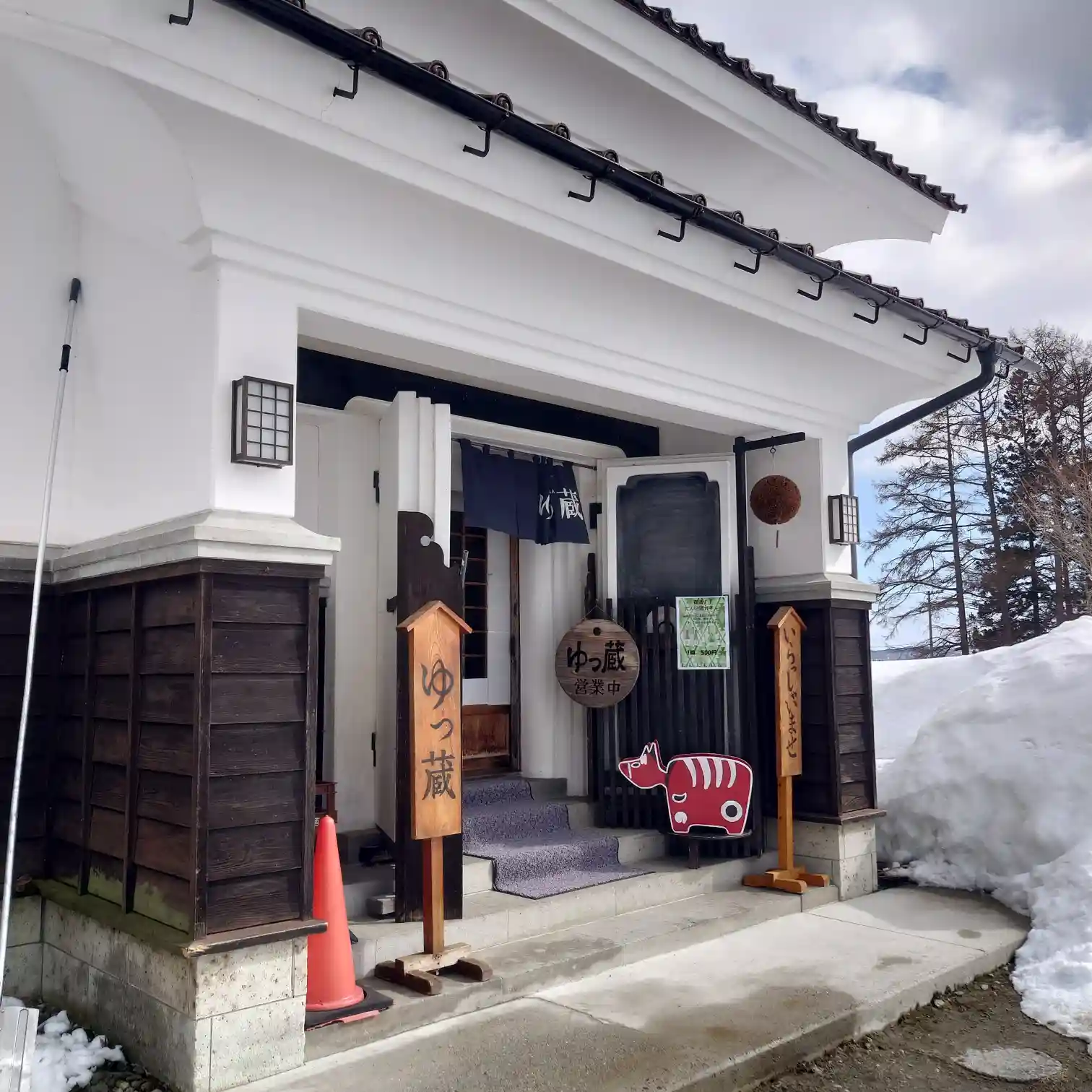 榮川酒造 ゆっ蔵の入口外観｜のれんと看板、赤べこが出迎える残雪期のエントランス