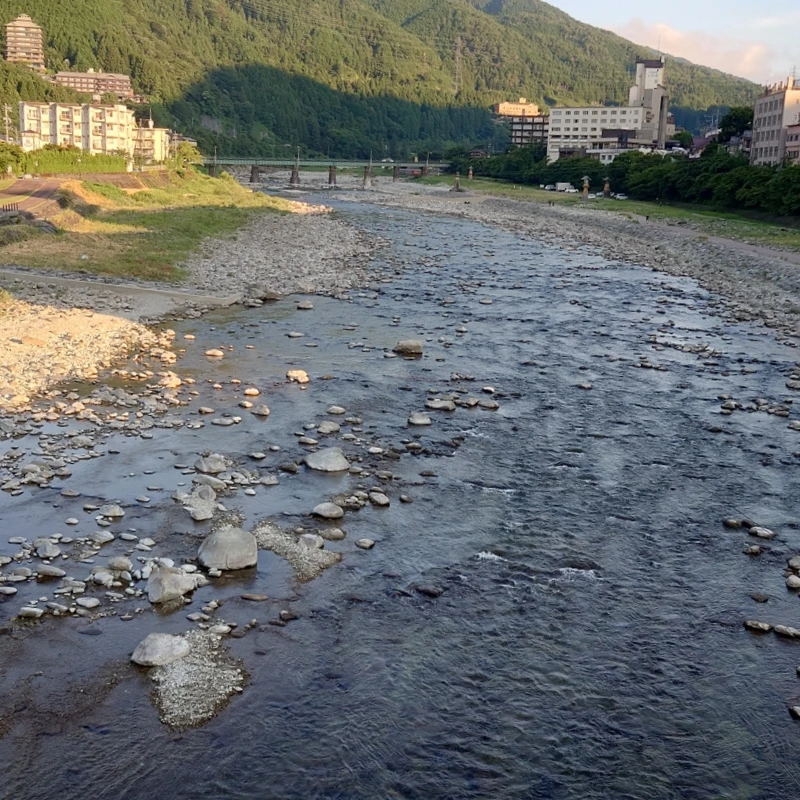 下呂温泉・飛騨川を橋上から望む清流風景。浅瀬に丸石が点在し、河川敷遊歩道と温泉街のホテル群、朝日に染まる山並みが広がるランコースの眺め