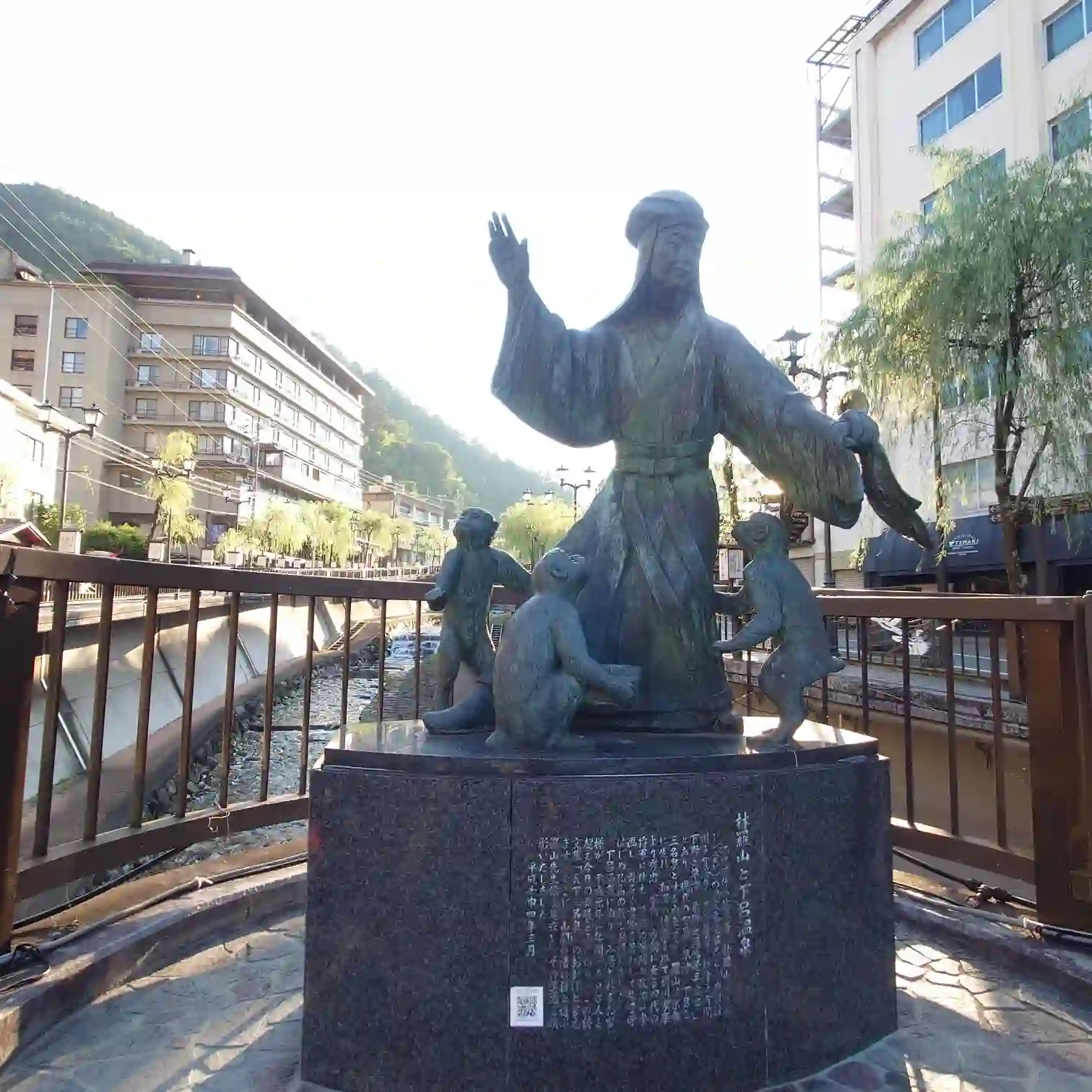 阿多野川に架かる橋のモニュメント。人物像と三匹の猿のブロンズ群像を近景で、背後に温泉街と柳並木が続く—菊半旅館発3kmコース
