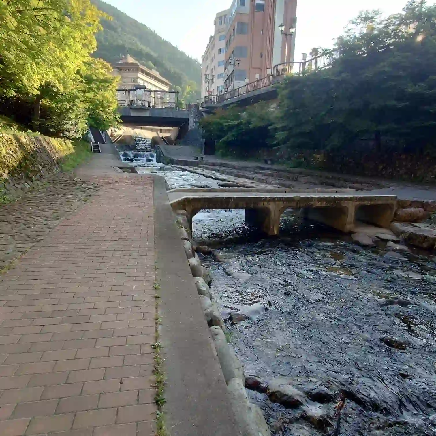 阿多野川沿いのレンガ遊歩道と低いコンクリ橋、前方に階段状の落ち込みと温泉街の建物—菊半旅館へ向かう復路/下呂温泉3kmコース