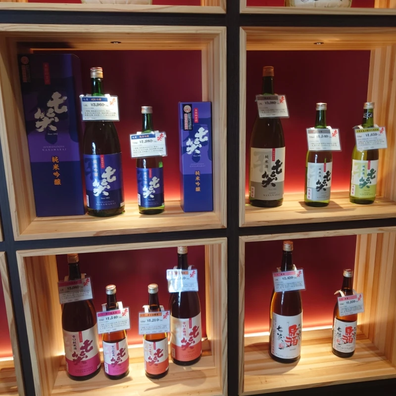 七笑酒造の店内棚。赤いバックライトの木製キューブに、純米吟醸〈紫箱〉や緑ラベル、一升瓶・四合瓶、桃・橙ラベルの銘柄が価格札付きで整然と並ぶ