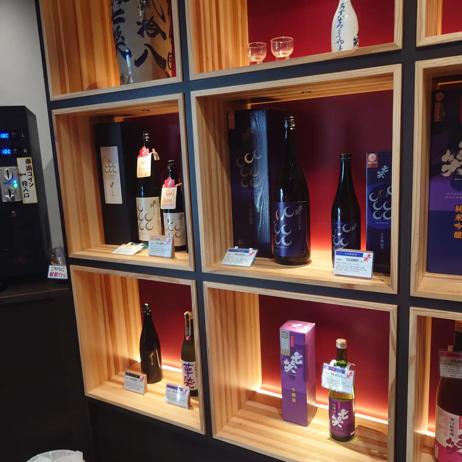 七笑酒造の店内ディスプレイ。赤いバックライトの木製キューブ棚に、月モチーフのラベルや紫箱の純米吟醸などが価格札付きで並び、上段にはお猪口も展示