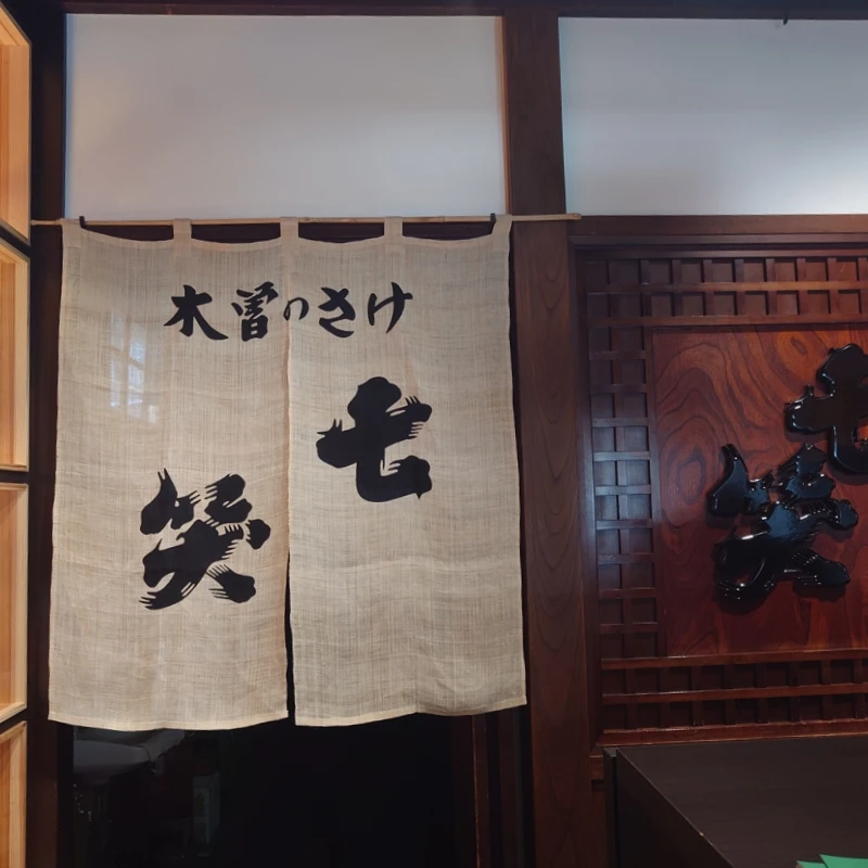 店内の麻のれんに『木曽のさけ 七笑』の筆文字。木格子と木目の壁、黒艶の立体ロゴが映える入口付近