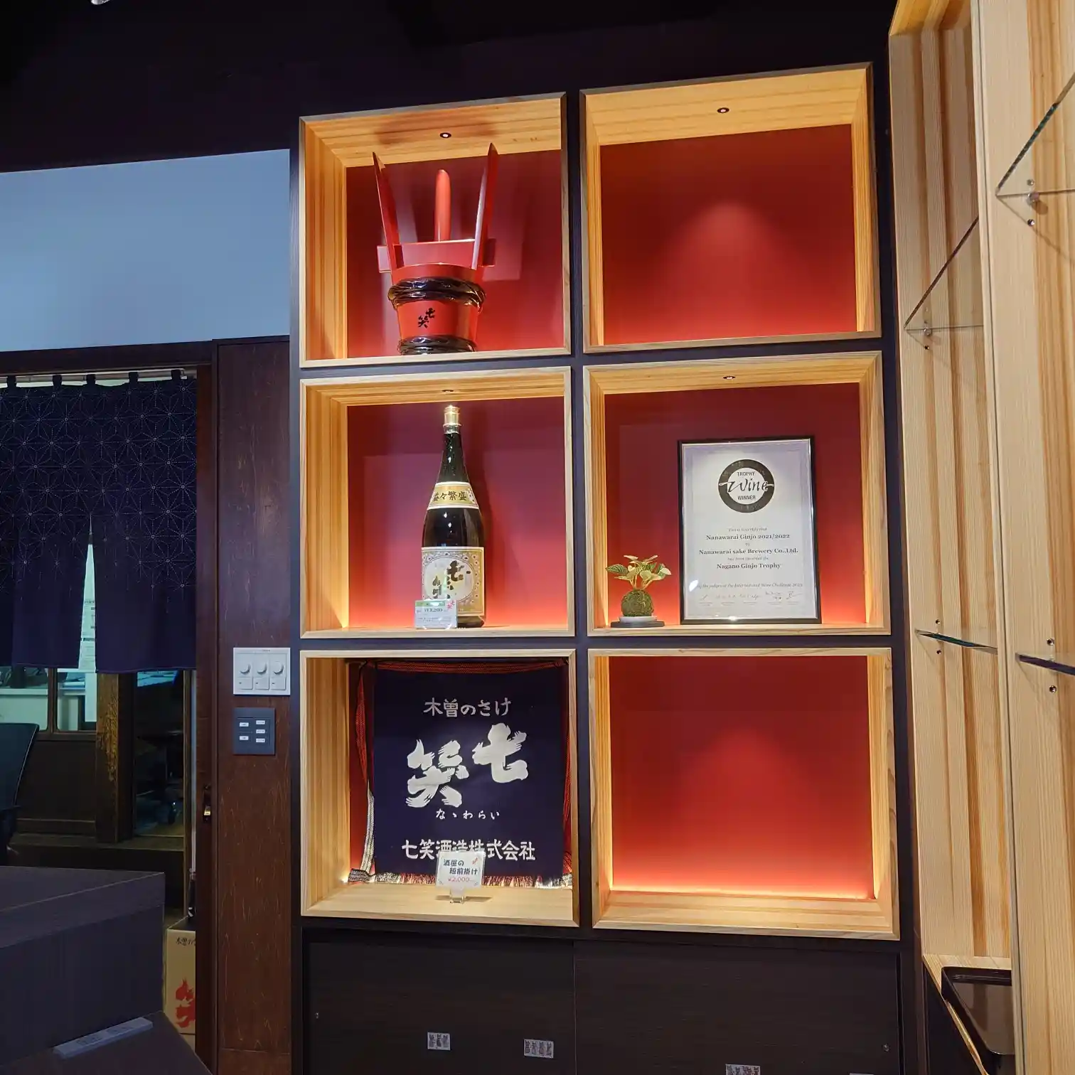 七笑酒造エントランス展示。赤いバックライトの木製キューブ棚に一升瓶、受賞トロフィーと賞状、蔵名入り前掛けや苔玉を配したディスプレイ。左手に紺ののれん
