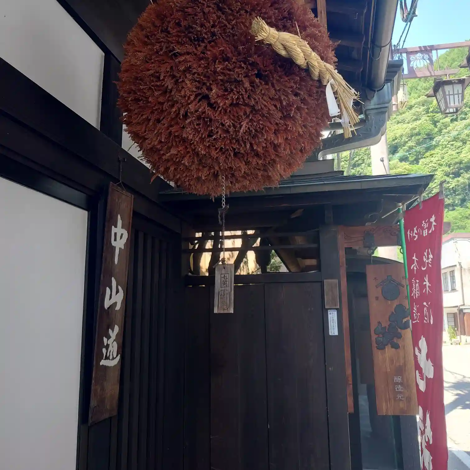 七笑酒造の軒先に吊るされた大ぶりの杉玉。『中山道』の木札と赤い幟が並び、奥に古い町並みと山の緑がのぞく入口風景