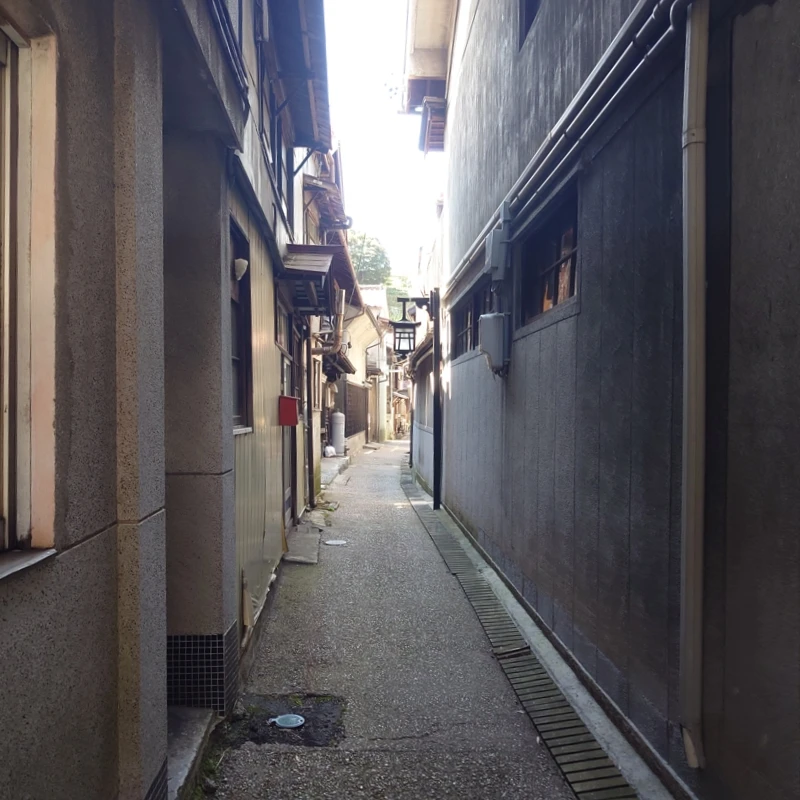 蔵の横を抜ける細い路地。両側を古い家並みが挟み、配管や行灯を横目に石舗装の小径が奥へまっすぐ伸びる