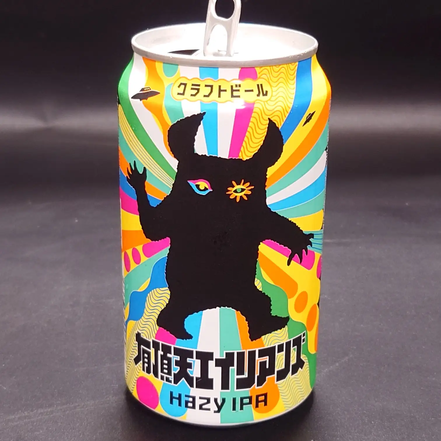 ヤッホーブルーイング『有頂天エイリアンズ HAZY IPA』の缶ビール。カラフルな放射状デザインに黒いエイリアンのシルエットと「クラフトビール」表記、黒背景で撮影。