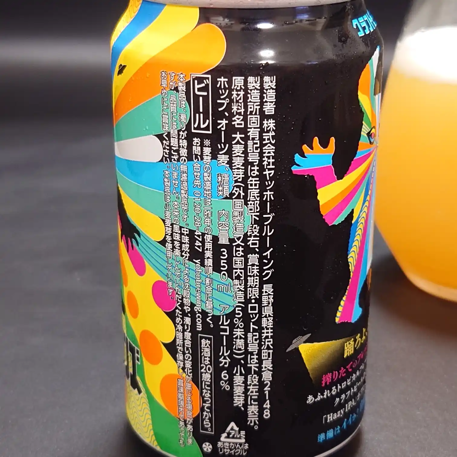 ヤッホーブルーイング『有頂天エイリアンズ HAZY IPA』缶の側面ラベル。原材料や製造者情報の表示をクローズアップ、右奥に注いだビールの濁ったオレンジ色がぼけて写る。