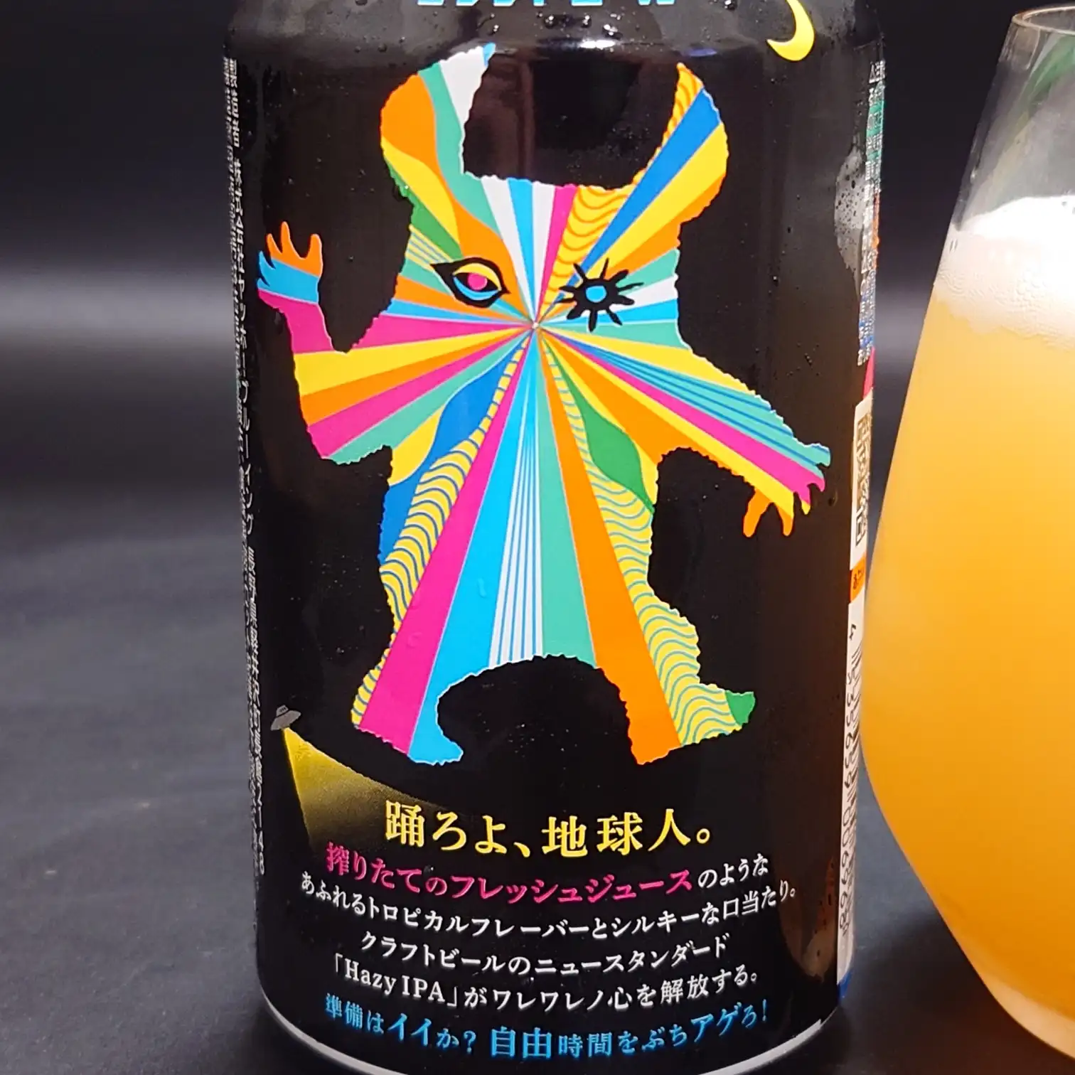 ヤッホーブルーイング『有頂天エイリアンズ HAZY IPA』缶の裏面デザイン接写。放射状カラーのエイリアンと「踊ろよ、地球人。」のコピー、右にグラスの濁りオレンジ色のビール。