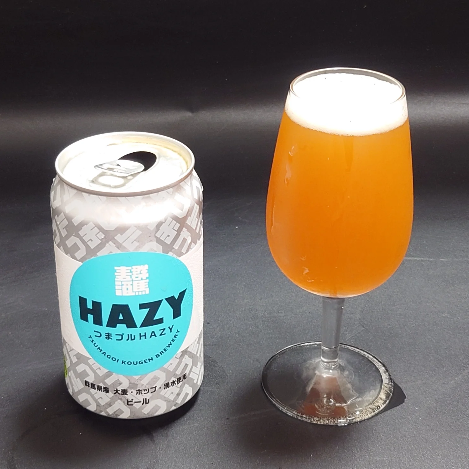 つまブル HAZYの缶とグラスのツーショット。濁りの強いオレンジ色の液体に白い泡、ターコイズのHAZYラベル缶。
