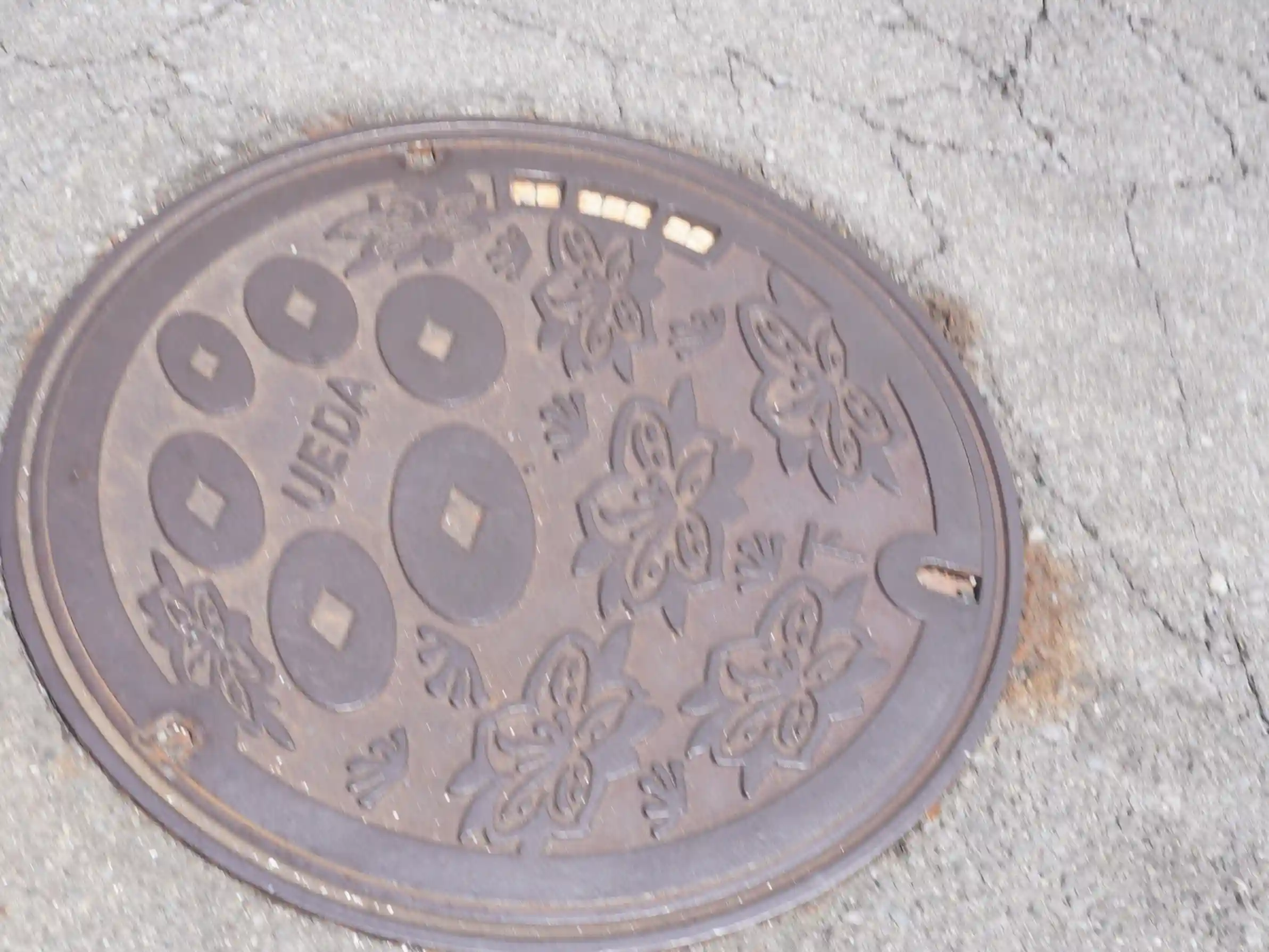 上田市のロゴ「UEDA」が入ったマンホール蓋。古銭と花の意匠（柳町通り近く）