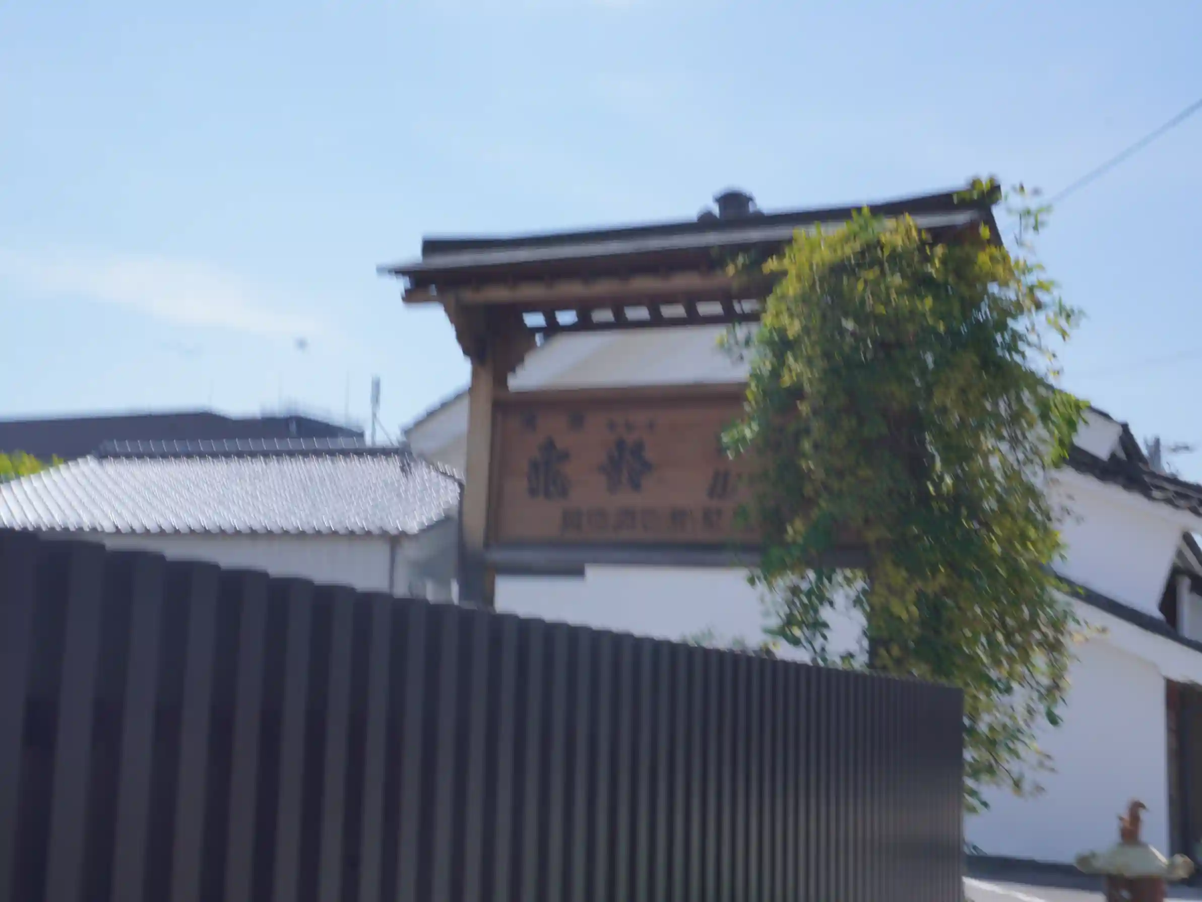 岡崎酒造の門に掲げられた木製の社名看板と白壁の蔵。蔦の緑がからむ外観（遠景）