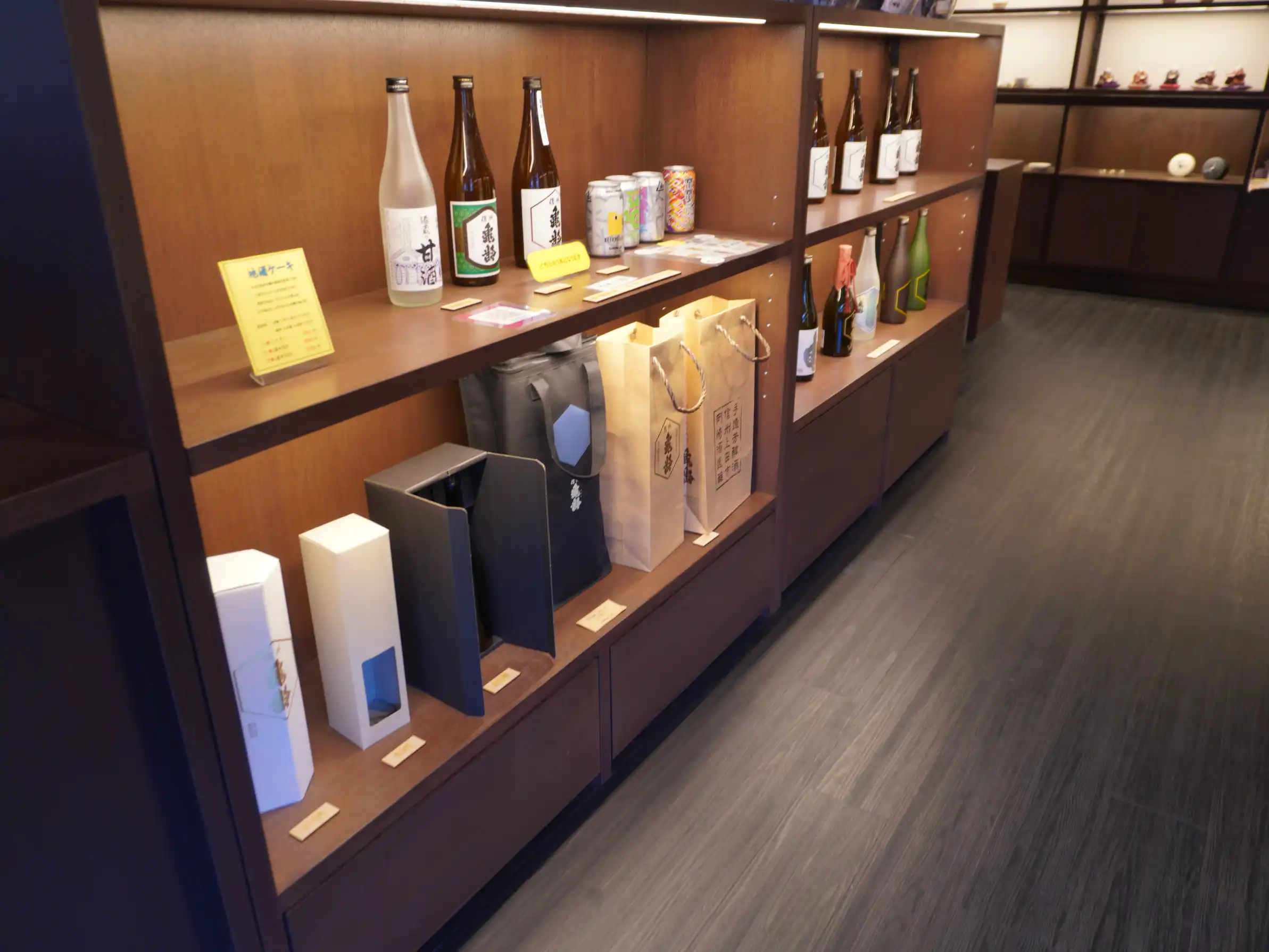 岡崎酒造の売店棚のロングショット。甘酒と信州亀齢の瓶、缶ビール、下段にギフト箱や手提げが並ぶ