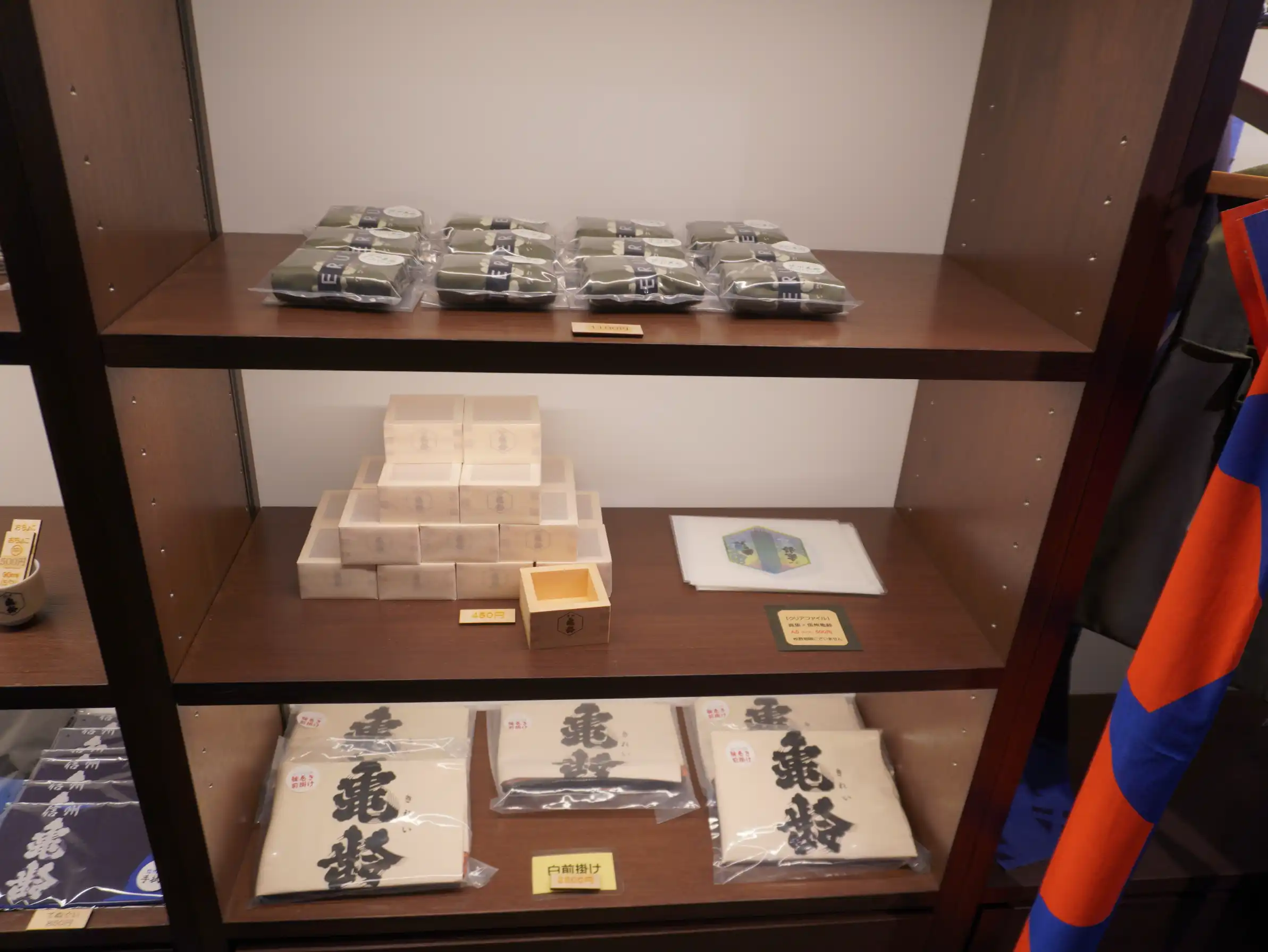 岡崎酒造の物販棚。亀齢ロゴの前掛けや手ぬぐい、木枡、雑貨が並ぶ（在庫・価格は変動）