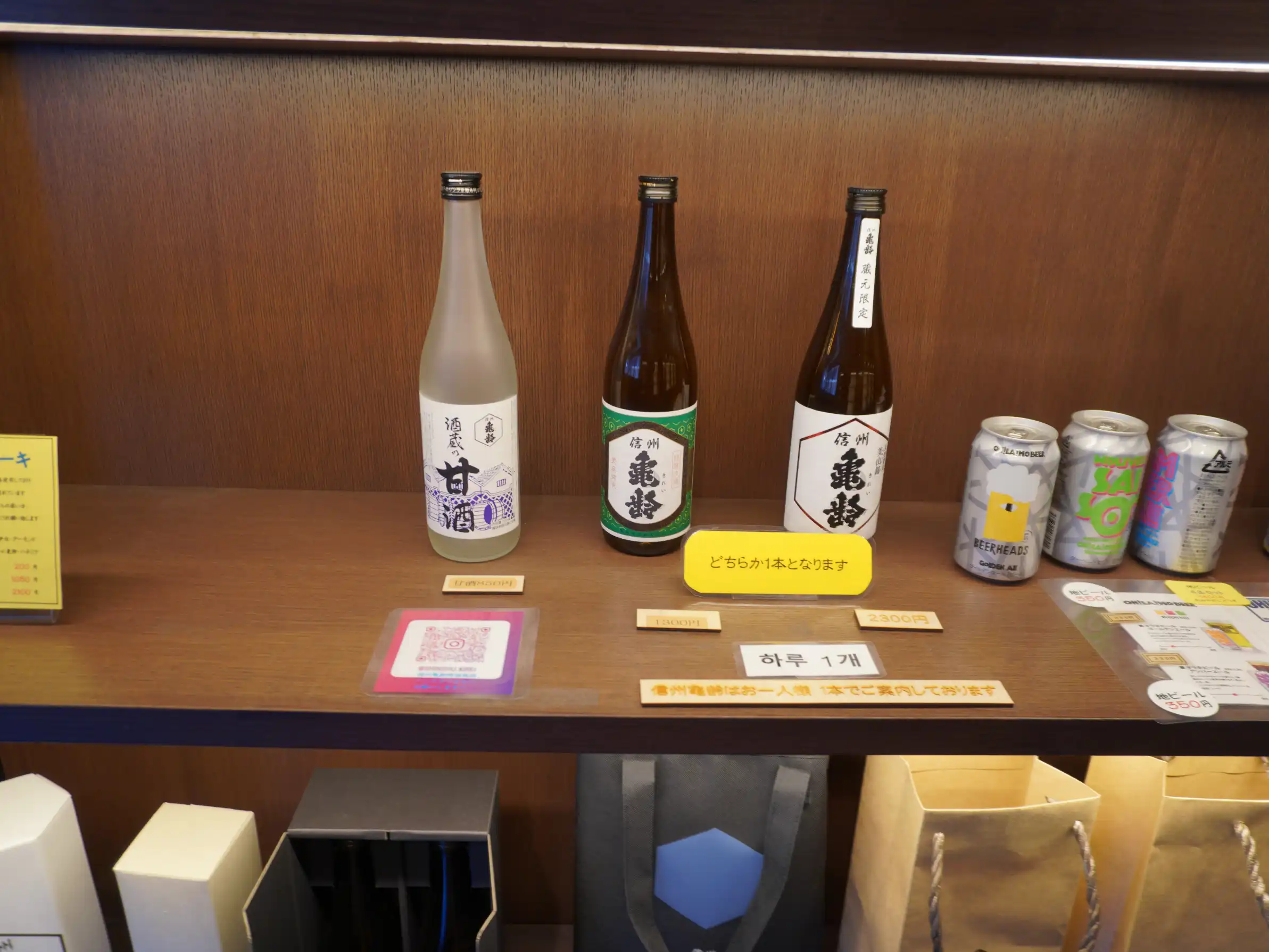 岡崎酒造の店頭棚に並ぶ本日の販売ラインナップ。信州亀齢の瓶（2種）と甘酒、右側に缶ビール。中央の札に「どちらか1本となります」案内