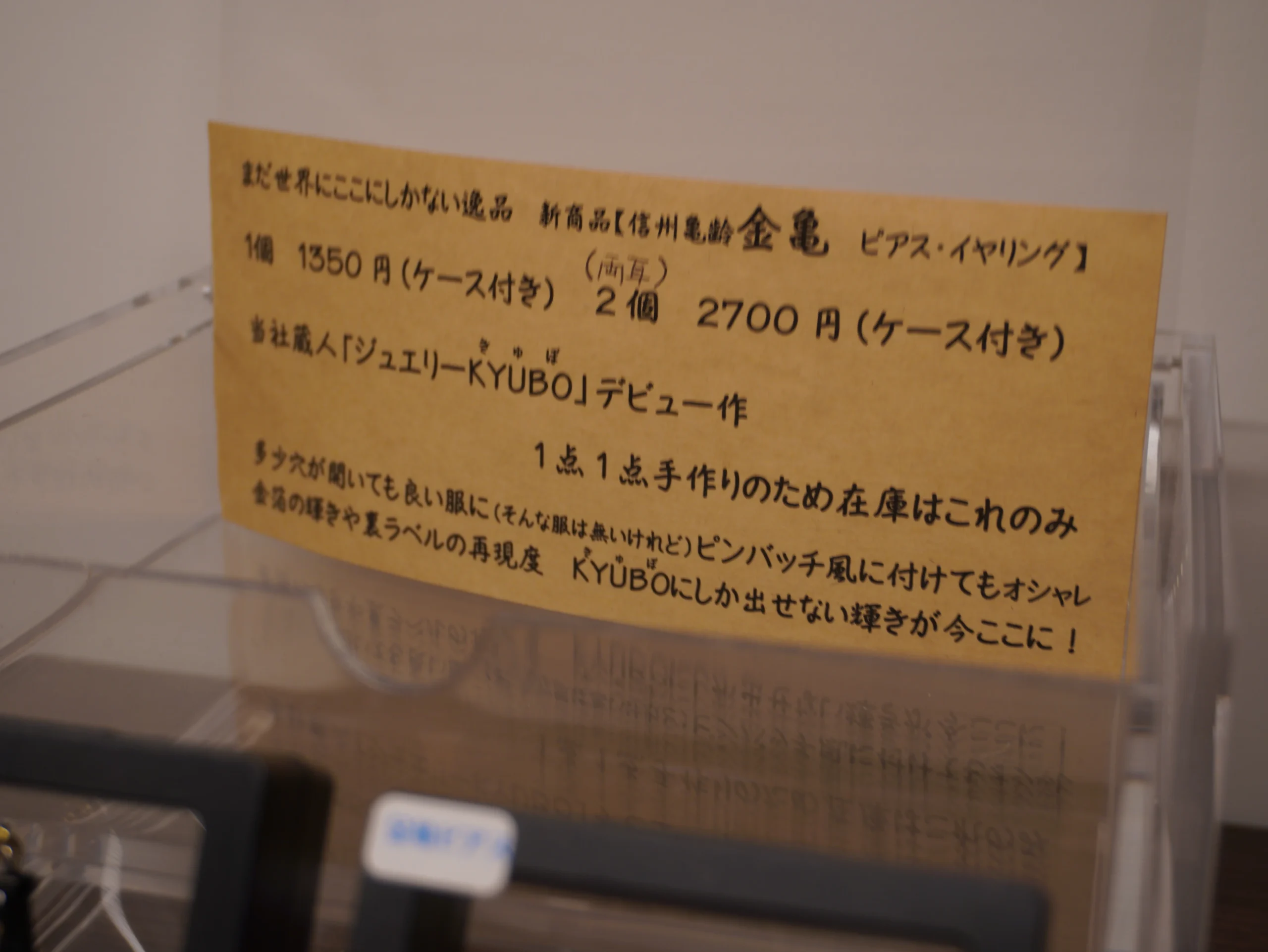 岡崎酒造の蔵人「ジュエリーKYUBO」デビュー作の酒瓶ピアス／イヤリングの案内札。価格例「1個1350円・2個2700円（ケース付き）」と製作表記