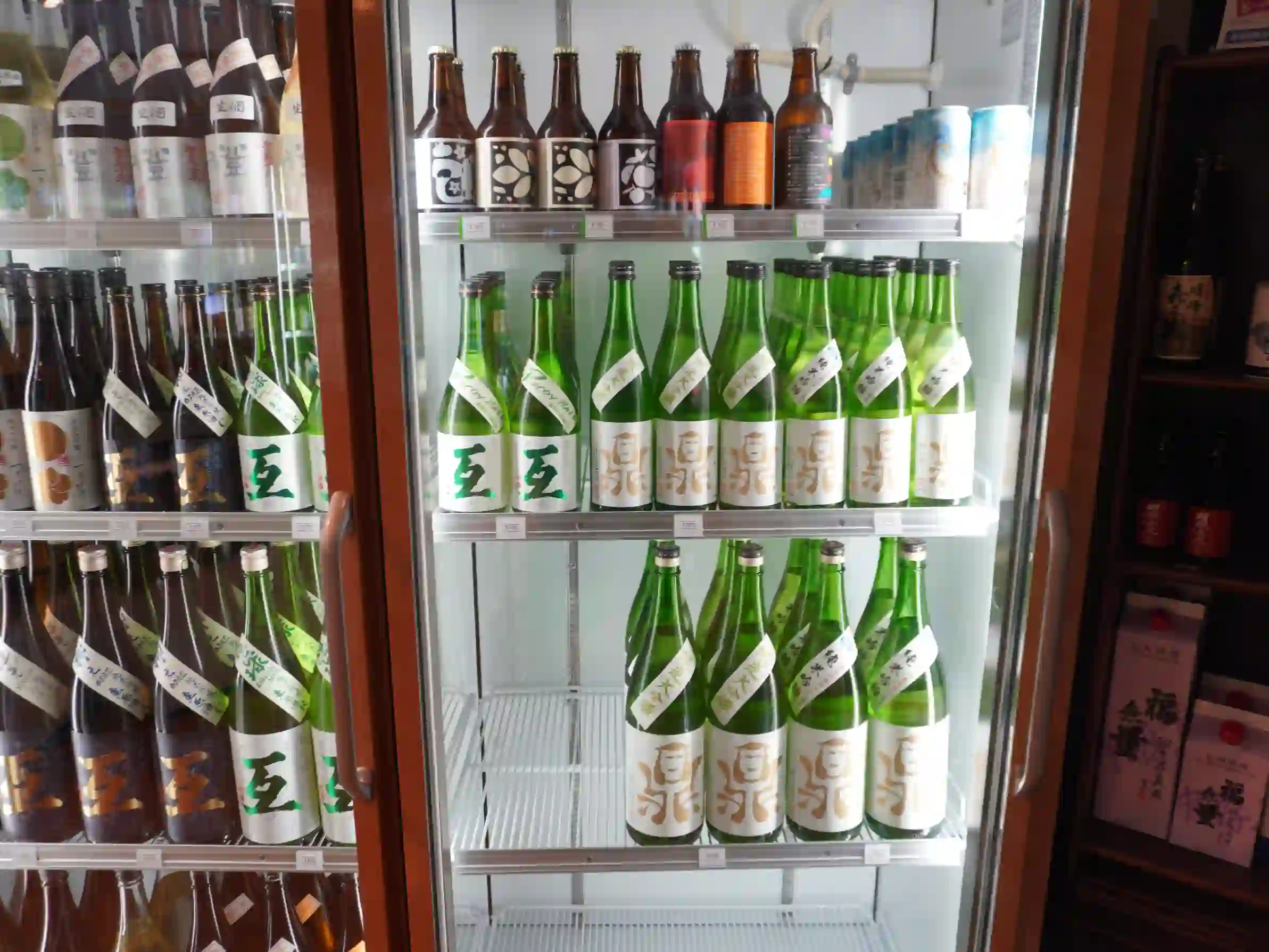 緑瓶の日本酒が並ぶ冷蔵棚。上段にクラフトビールの瓶と缶。