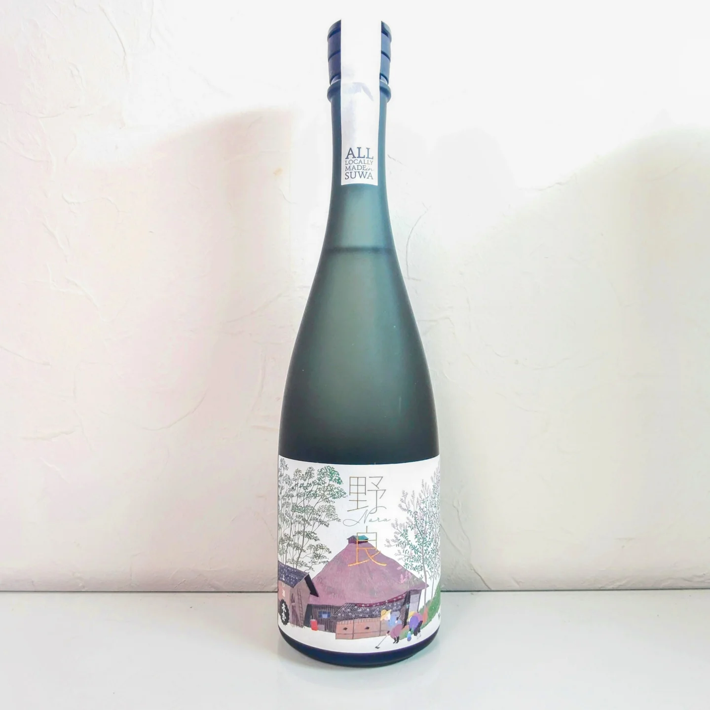 日本酒「野良 Nora」本金—すりガラス調の黒瓶に原田泰治のイラストラベルをまとった全体写真、美術品のような佇まい