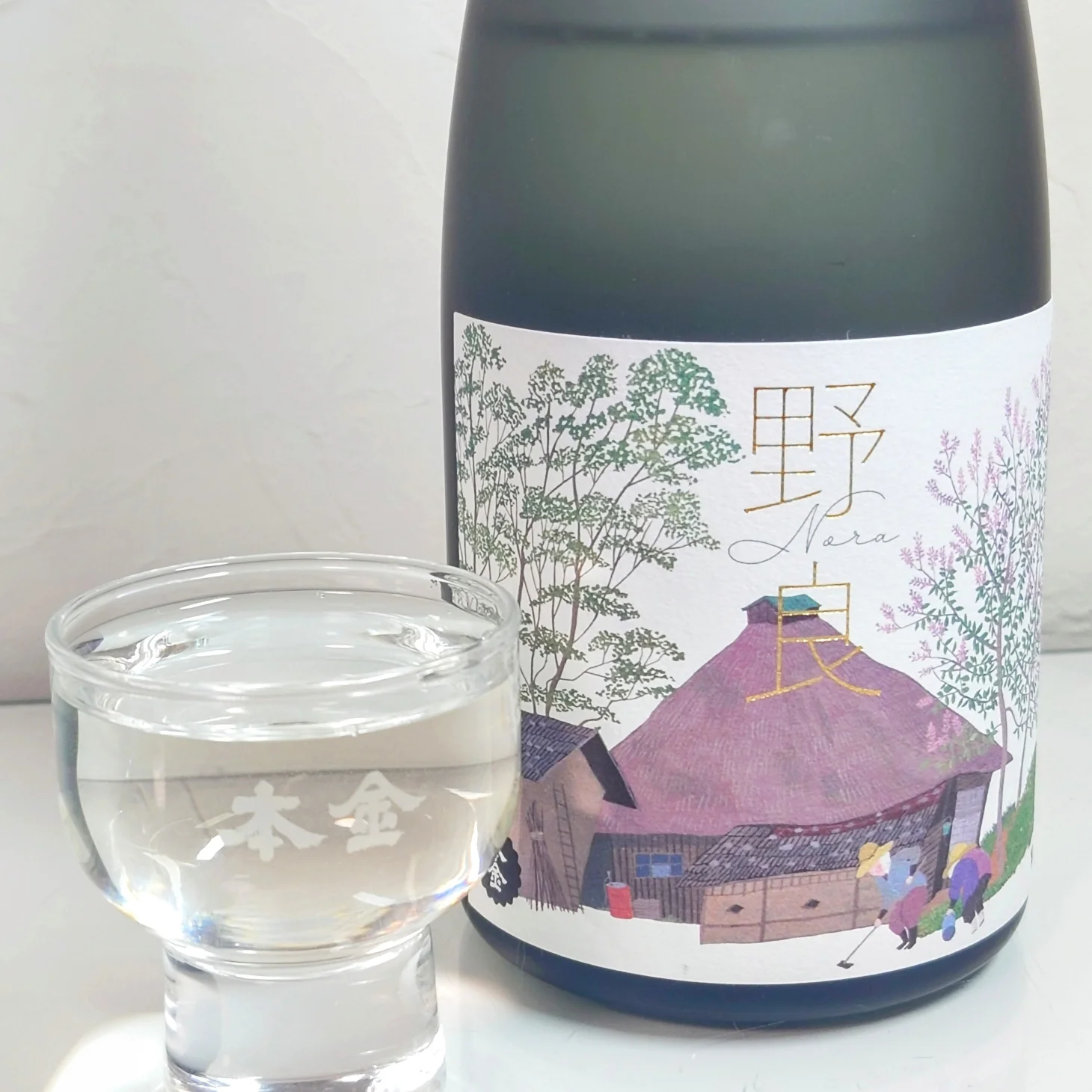 日本酒「野良 Nora」本金—原田泰治のイラストラベル瓶と本金銘入りの盃に注がれた透明な酒、試飲シーン