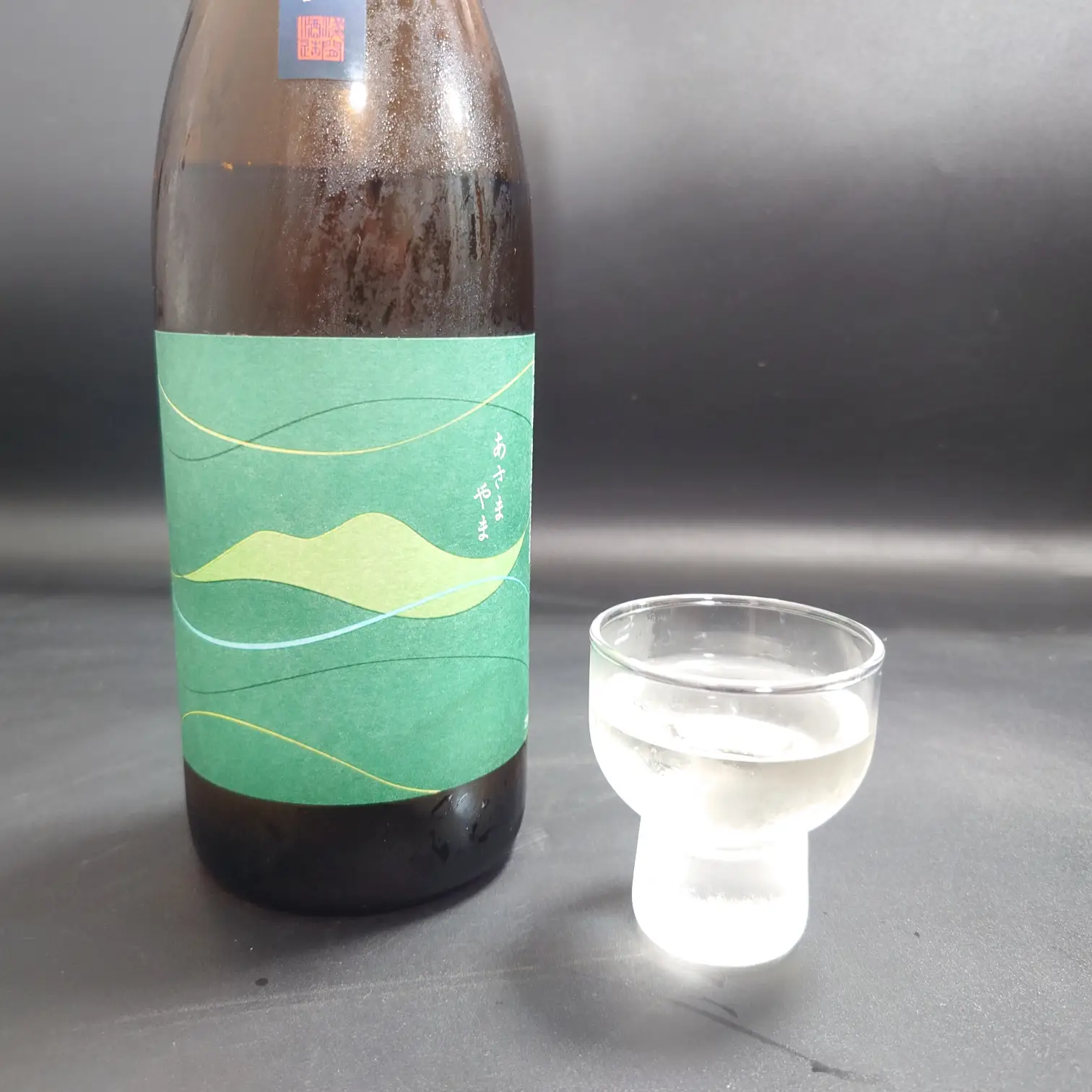 八ッ場ダム貯蔵酒|あさやまボトルとお猪口の並び 群馬・浅間酒造「八ッ場ダム貯蔵酒」あさやまのボトルと透明な酒を注いだガラスのお猪口を並べて撮影した写真