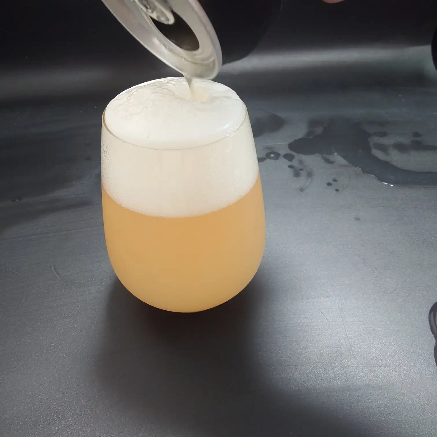 宇宙ビール 仕上げの注ぎ ヘイジーゴールドと厚い泡 UCHU BREWING 宇宙ビール ヘイジーな淡いゴールドをステムレスグラスにたっぷり注ぎ込み こんもりした厚い泡が盛り上がる瞬間 黒背景に水滴の跡が見える 山梨・北杜のクラフトビール