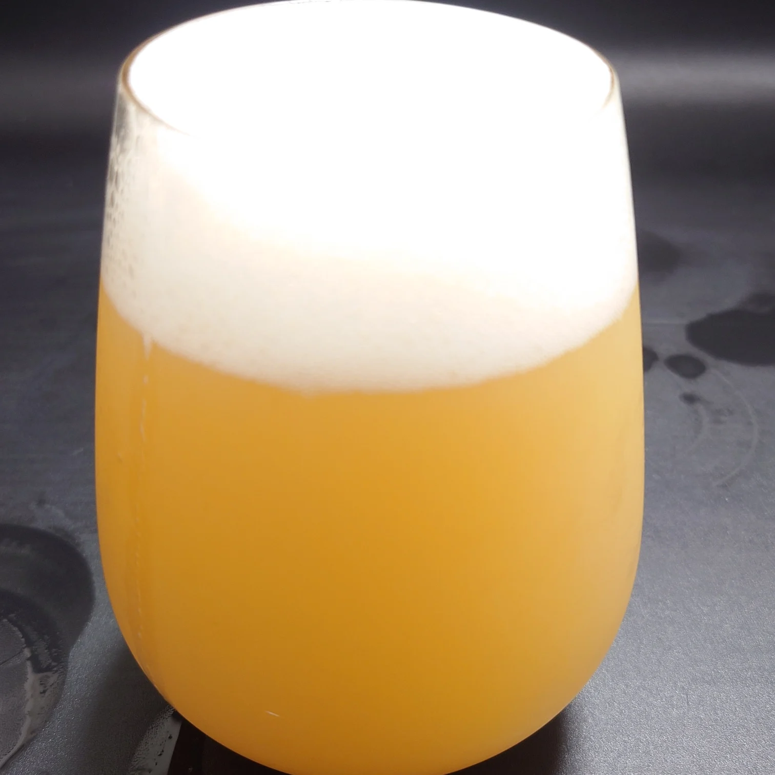 宇宙ビール 液色クローズアップ ヘイジーゴールドと厚い泡 UCHU BREWING 宇宙ビール ヘイジーな濁りの濃いゴールドときめ細かな白い泡の層をステムレスグラスでアップで切り取った 黒背景に水滴跡が見える