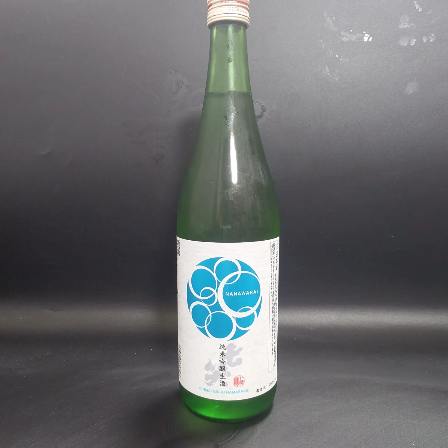七笑 純米吟醸生酒のボトル正面ラベル 青い円環ロゴNANAWARAI 黒背景で紙質のテクスチャーが分かる
