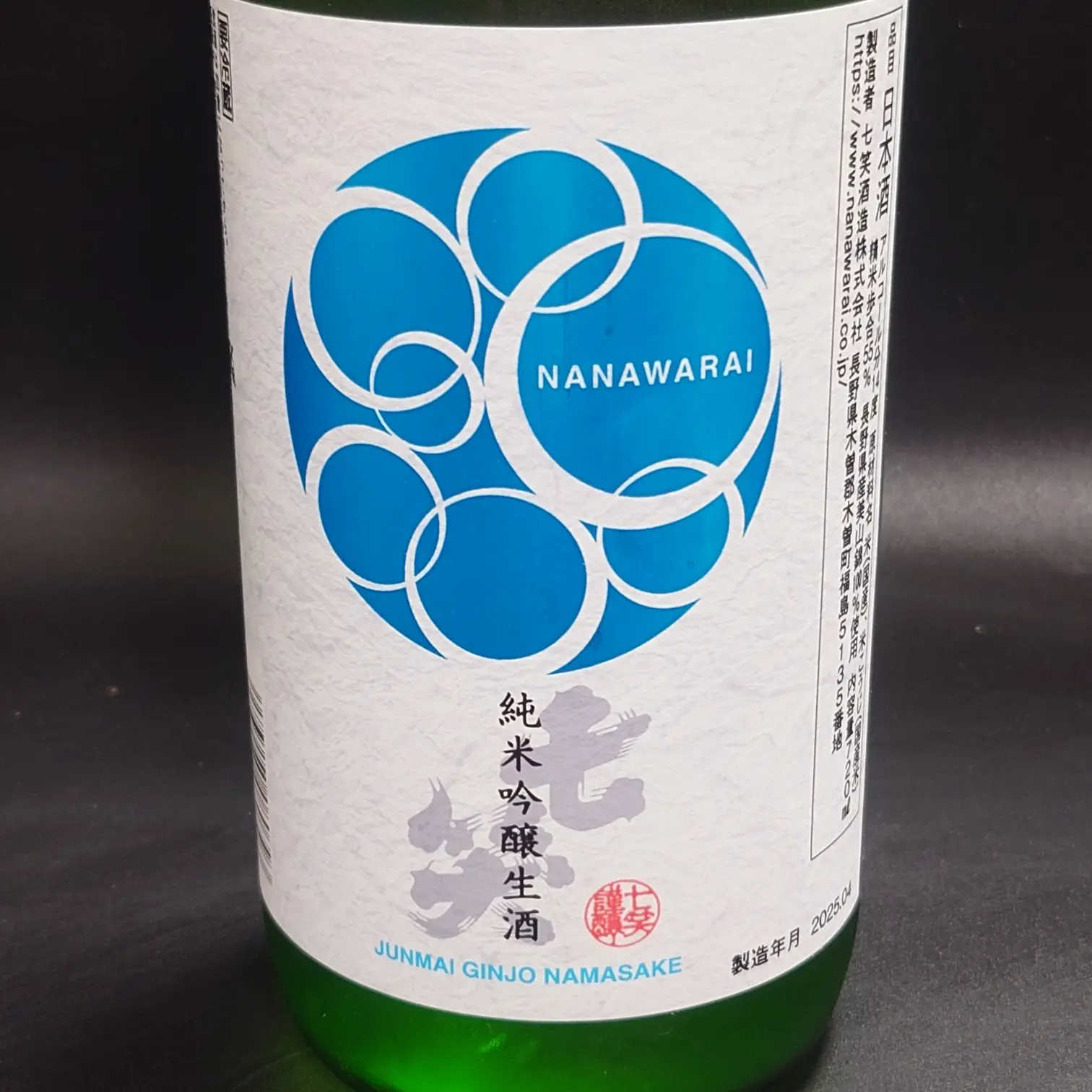 七笑 純米吟醸生酒のボトル正面ラベル 青い円環ロゴNANAWARAIが目立つ グリーンボトルのクローズアップ 黒背景で紙質のテクスチャーが分かる
