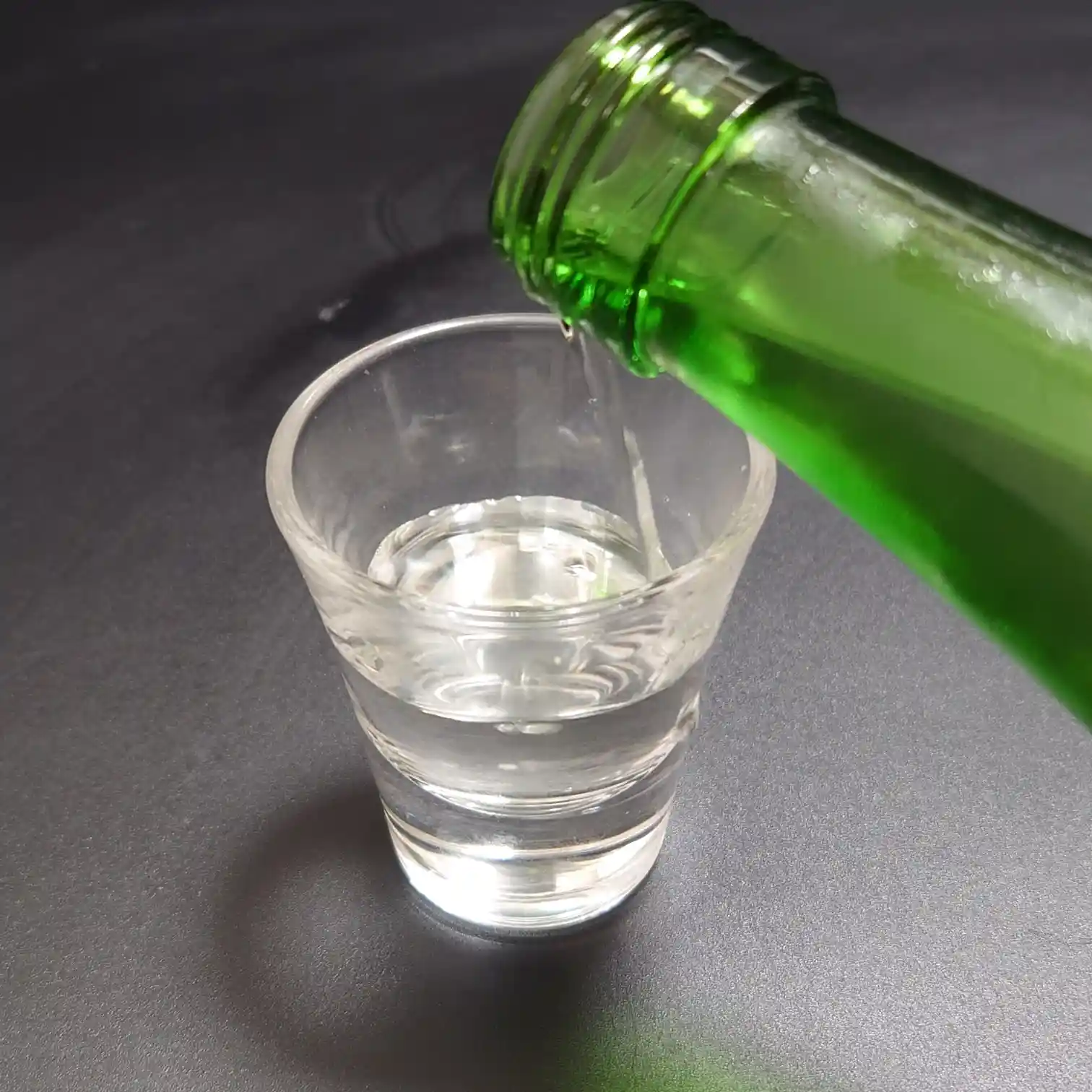 七笑 純米吟醸生酒をグリーンボトルからグラスへ注ぐ瞬間 透明感ある液体が滴り落ちるクローズアップ 黒背景で日本酒の澄んだ輝きが際立つ