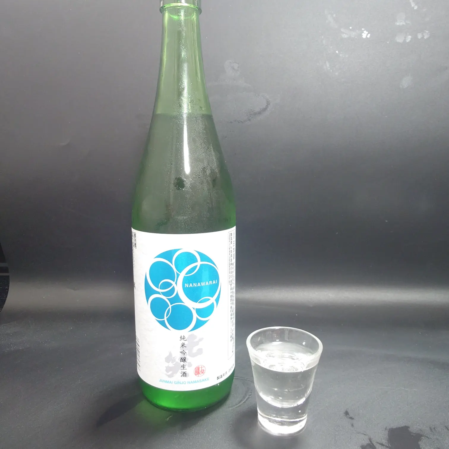 七笑 純米吟醸生酒のボトルとグラスショット 冷えた緑瓶の正面ラベルと透明感ある酒が入ったグラスを並べて撮影 黒背景に酒の清涼感が映える