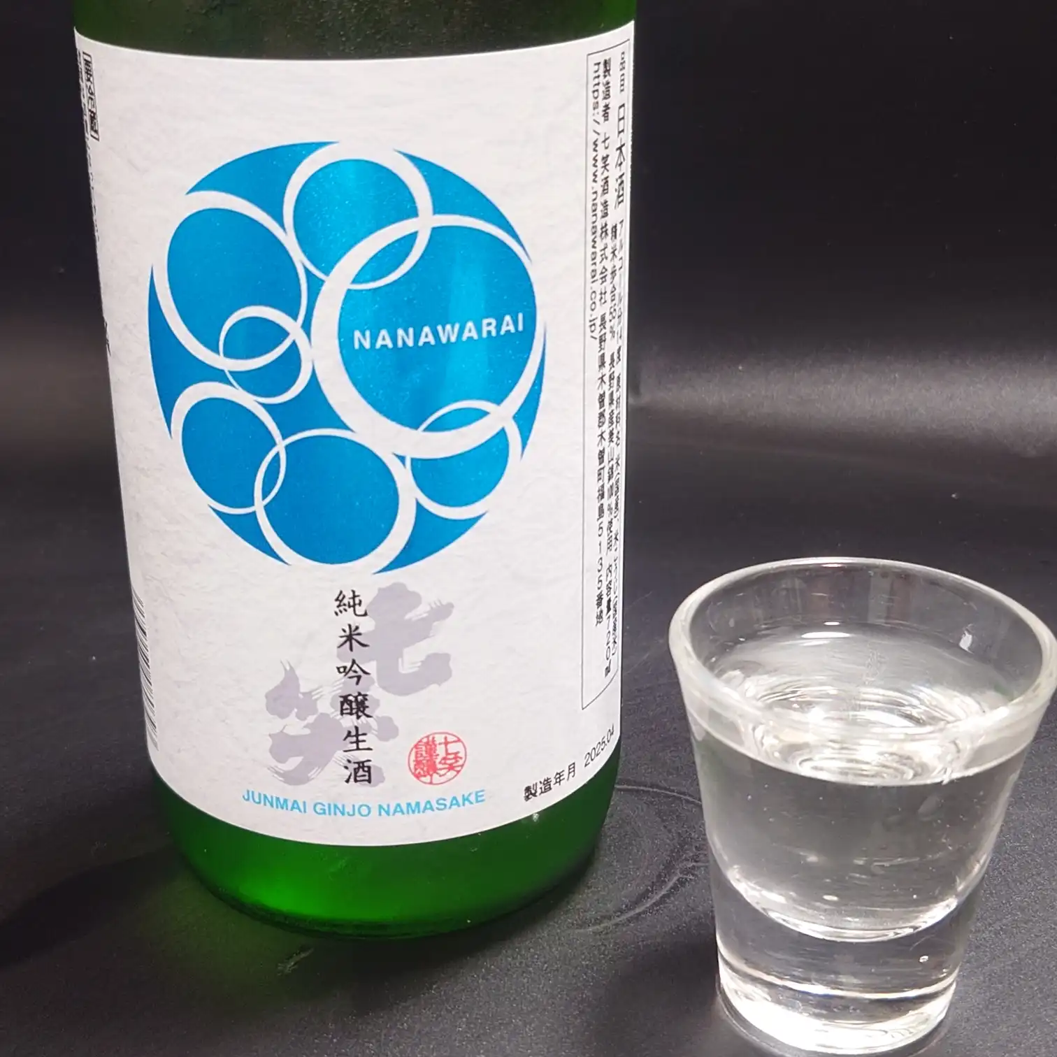 七笑 純米吟醸生酒の正面ラベルとグラスをクローズアップ 青い円環ロゴと純米吟醸生酒の文字が映え 透明な酒が注がれたグラスが横に並ぶ