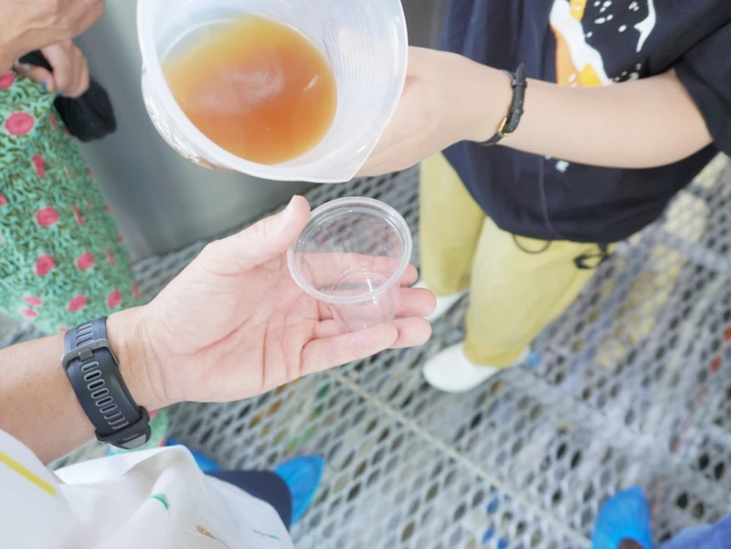 第2章 出来立ての麦汁を注ぐ|透明カップに広がる甘い香り 長野・佐久 ヤッホーブルーイング よなよなエールの醸造所見学 第2章|金属グレーチング上で透明カップに計量カップから出来立ての甘い麦汁を注ぐ瞬間が伝わる手元の一枚