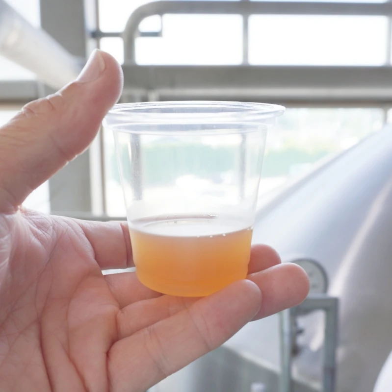 第2章 仕込み釜を背景に|透明カップの琥珀色の麦汁 長野・佐久 ヤッホーブルーイング よなよなエールの醸造所見学 第2章|仕込み釜を背景に注ぎたての琥珀色の麦汁を透明カップで掲げる手元 淡い泡と温かな色合いが伝わる