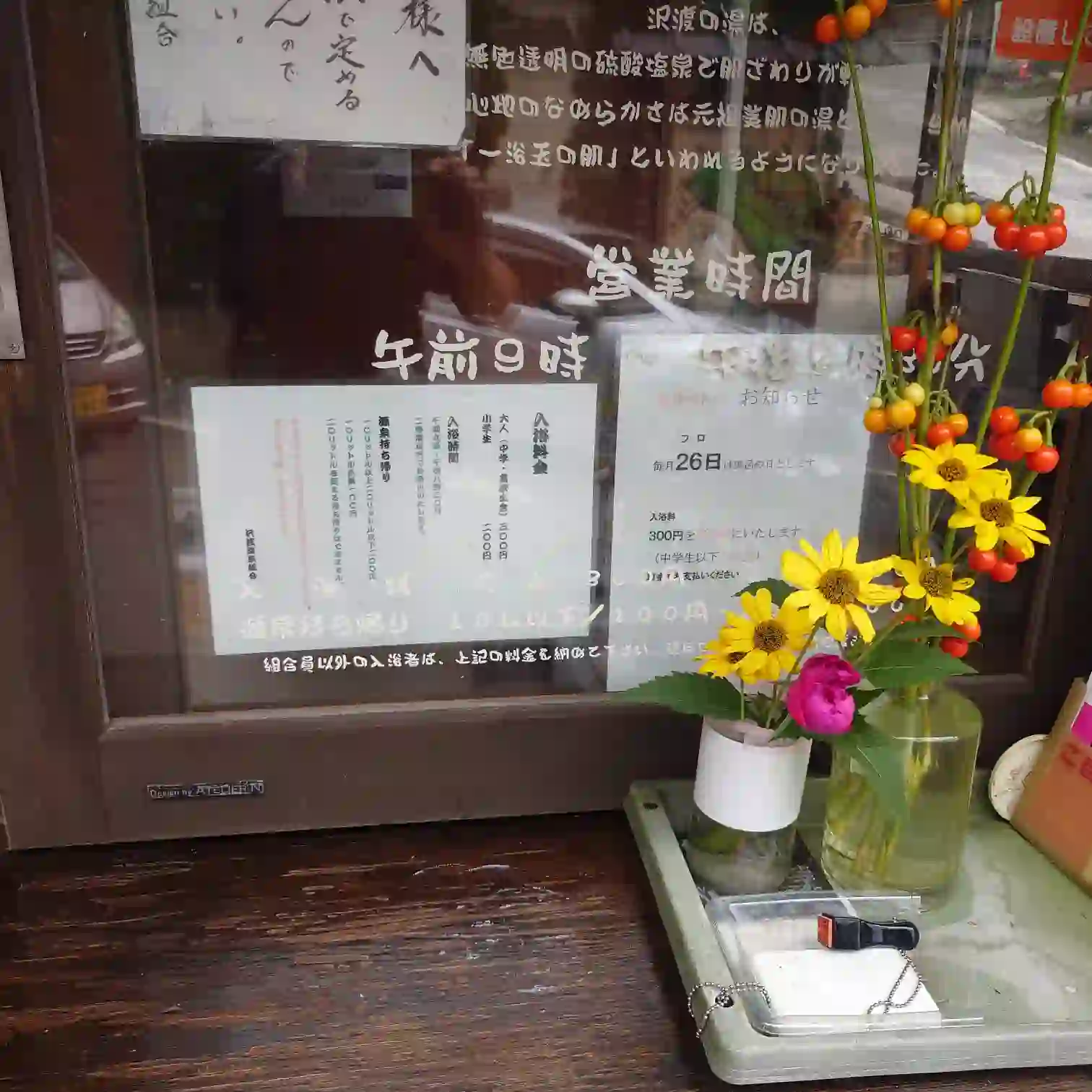 沢渡温泉共同浴場の受付窓口。営業時間や料金の掲示と、花瓶の黄色い花が出迎える。