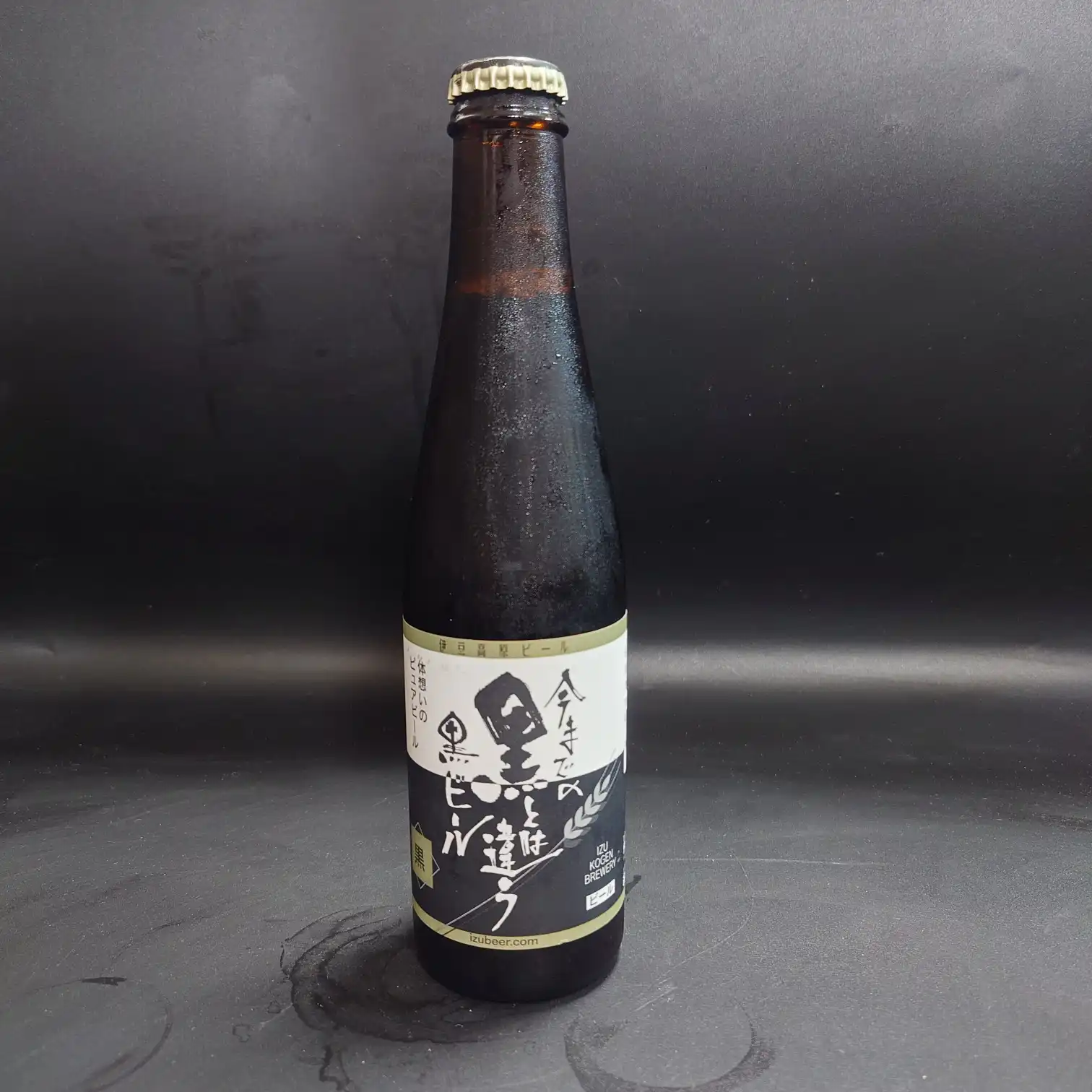 黒地背景に立てた小瓶の伊豆高原ビール黒スタウト 冷えたガラスに水滴がつき 白い筆文字のラベルが目立つ 正面ショット