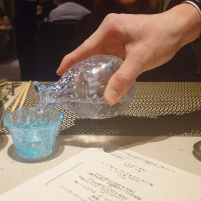 ルッチコーレでソムリエが新政を注ぐ所作（ガラス徳利から青いガラス盃へ、丁寧な手元の瞬間）