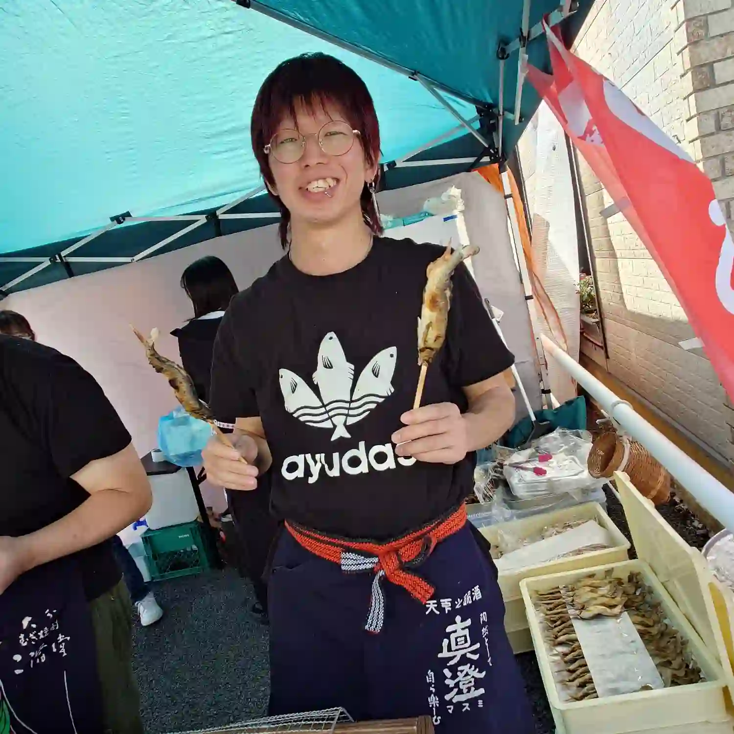ayudasのTシャツを着て両手に鮎を持つ青年